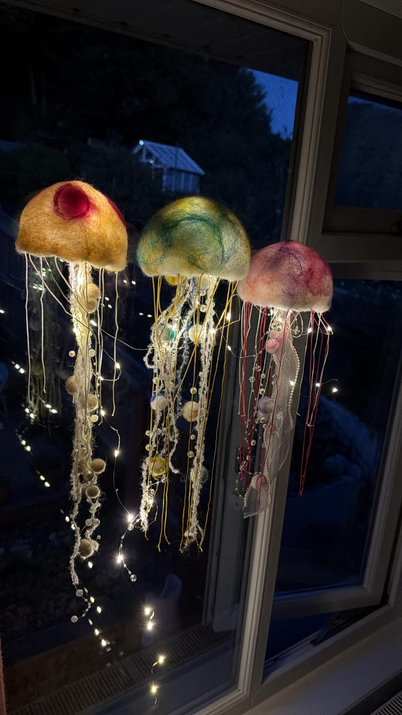 May include: Three hanging jellyfish decorations crafted from felted wool in yellow, green, and pink hues. Each jellyfish features long, dangling tentacles adorned with small beads and integrated warm white LED fairy lights that illuminate the structures.