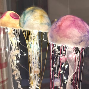 May include: Three hanging jellyfish ornaments crafted from soft, fuzzy material with rounded caps in shades of yellow, blue, and pink. Each jellyfish features long, dangling tentacles made of thin strings, beads, and integrated glowing LED lights.
