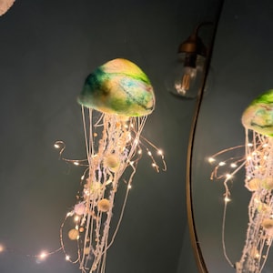 May include: A hanging jellyfish decoration featuring a felted dome in shades of green, blue, and yellow. The structure includes long, dangling white fibers and thin copper wire strands illuminated by small, warm white LED fairy lights.