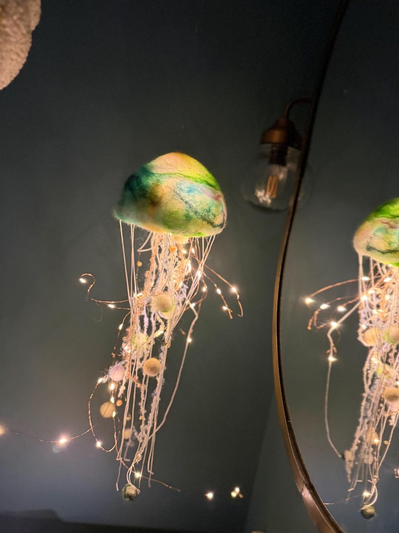 May include: A hanging jellyfish decoration featuring a felted dome in shades of green, blue, and yellow. The structure includes long, dangling white fibers and thin copper wire strands illuminated by small, warm white LED fairy lights.
