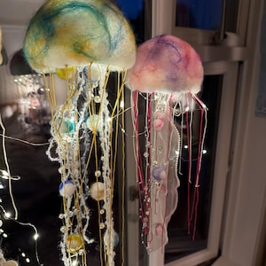 May include: Two hanging jellyfish ornaments crafted from felted wool. The left jellyfish features a teal and yellow dome with yellow threads and pearl beads. The right jellyfish has a pink and purple dome with red threads and white lace accents.