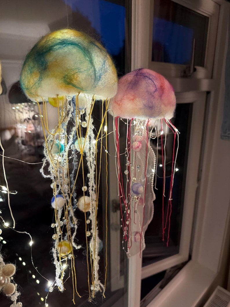May include: Two hanging jellyfish ornaments crafted from felted wool. The left jellyfish features a teal and yellow dome with yellow threads and pearl beads. The right jellyfish has a pink and purple dome with red threads and white lace accents.