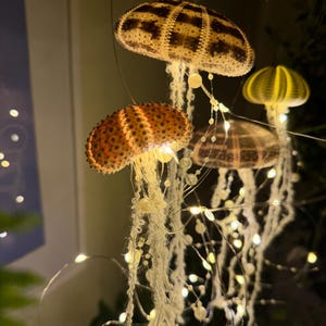 Peut inclure: Suspensions décoratives en forme de méduses. Les lumières présentent un motif texturé et tacheté dans des tons marron et jaune. Les méduses ont de longs tentacules blancs en forme de ficelle et sont éclairées par de petites lumières LED chaudes.