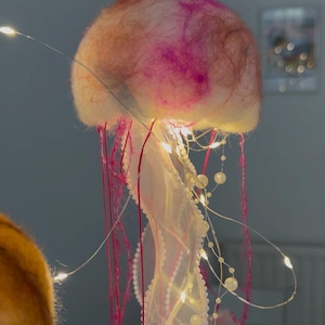 May include: A jellyfish-shaped hanging decoration featuring a soft, rounded top made of felted wool in shades of pink, orange, and white. It includes long, thin red threads, white mesh ribbons, and strings of small white beads illuminated by tiny LED lights.