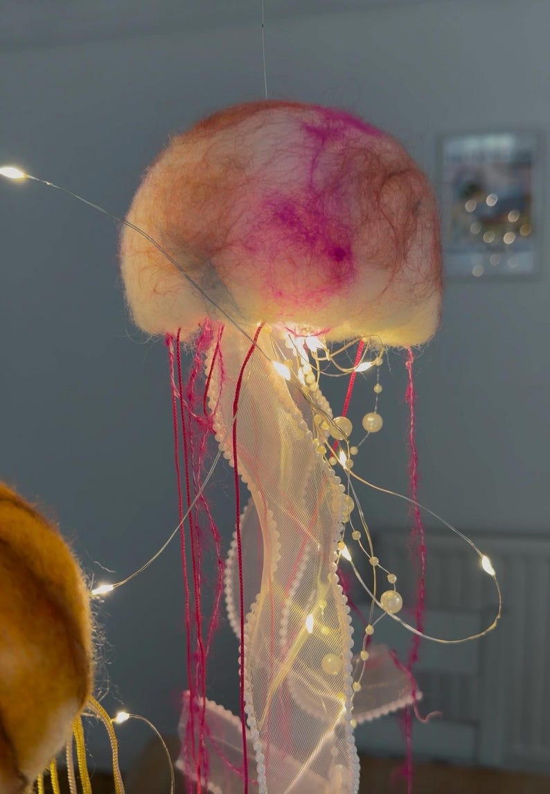 May include: A jellyfish-shaped hanging decoration featuring a soft, rounded top made of felted wool in shades of pink, orange, and white. It includes long, thin red threads, white mesh ribbons, and strings of small white beads illuminated by tiny LED lights.