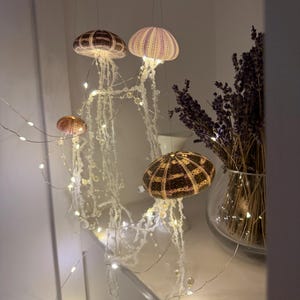 Op de afbeelding: Decoratieve hanglampen in de vorm van kwallen met ingewikkelde patronen en warme verlichting. De kwallen hangen aan het plafond en creëren een grillige en artistieke weergave. Een vaas met lavendel voegt een natuurlijk tintje toe.