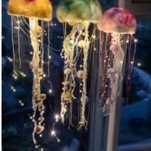 May include: Three hanging jellyfish decorations crafted from felted wool in yellow, green, and pink hues. Each jellyfish features long, dangling tentacles adorned with small beads and integrated warm white LED fairy lights that illuminate the structures.