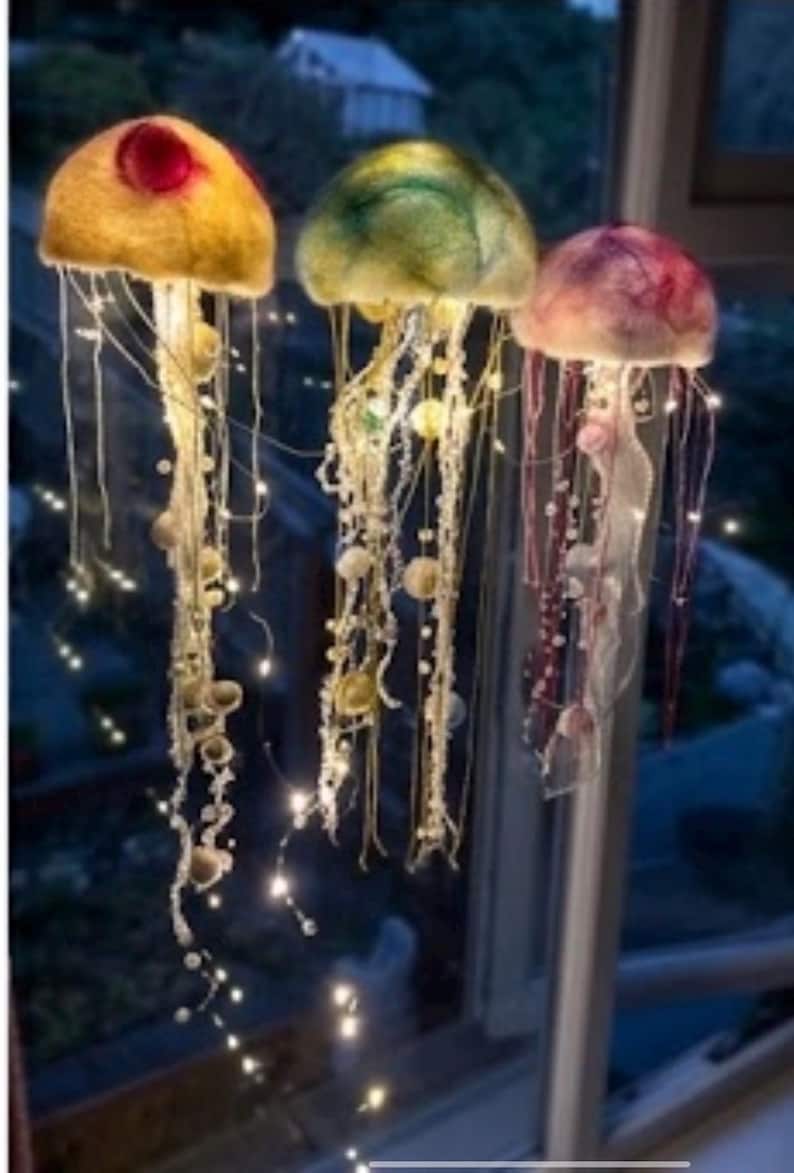 May include: Three hanging jellyfish decorations crafted from felted wool in yellow, green, and pink hues. Each jellyfish features long, dangling tentacles adorned with small beads and integrated warm white LED fairy lights that illuminate the structures.
