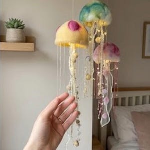 May include: A hanging mobile featuring three jellyfish-shaped ornaments made of felted wool. The jellyfish are yellow, teal, and pink, each adorned with long, dangling beaded tentacles and integrated small lights that emit a soft glow.
