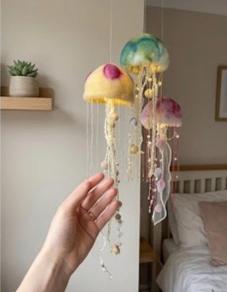 May include: A hanging mobile featuring three jellyfish-shaped ornaments made of felted wool. The jellyfish are yellow, teal, and pink, each adorned with long, dangling beaded tentacles and integrated small lights that emit a soft glow.