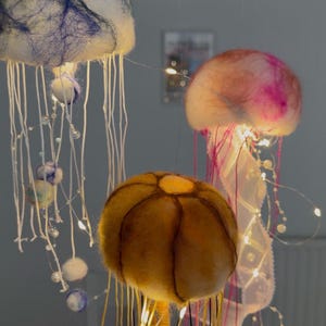 May include: Three hanging jellyfish decorations made of felted wool. One is white with blue fibers, one is pink and white, and one is golden yellow with dark brown segment lines. Each features long dangling white and colored threads and small LED lights.