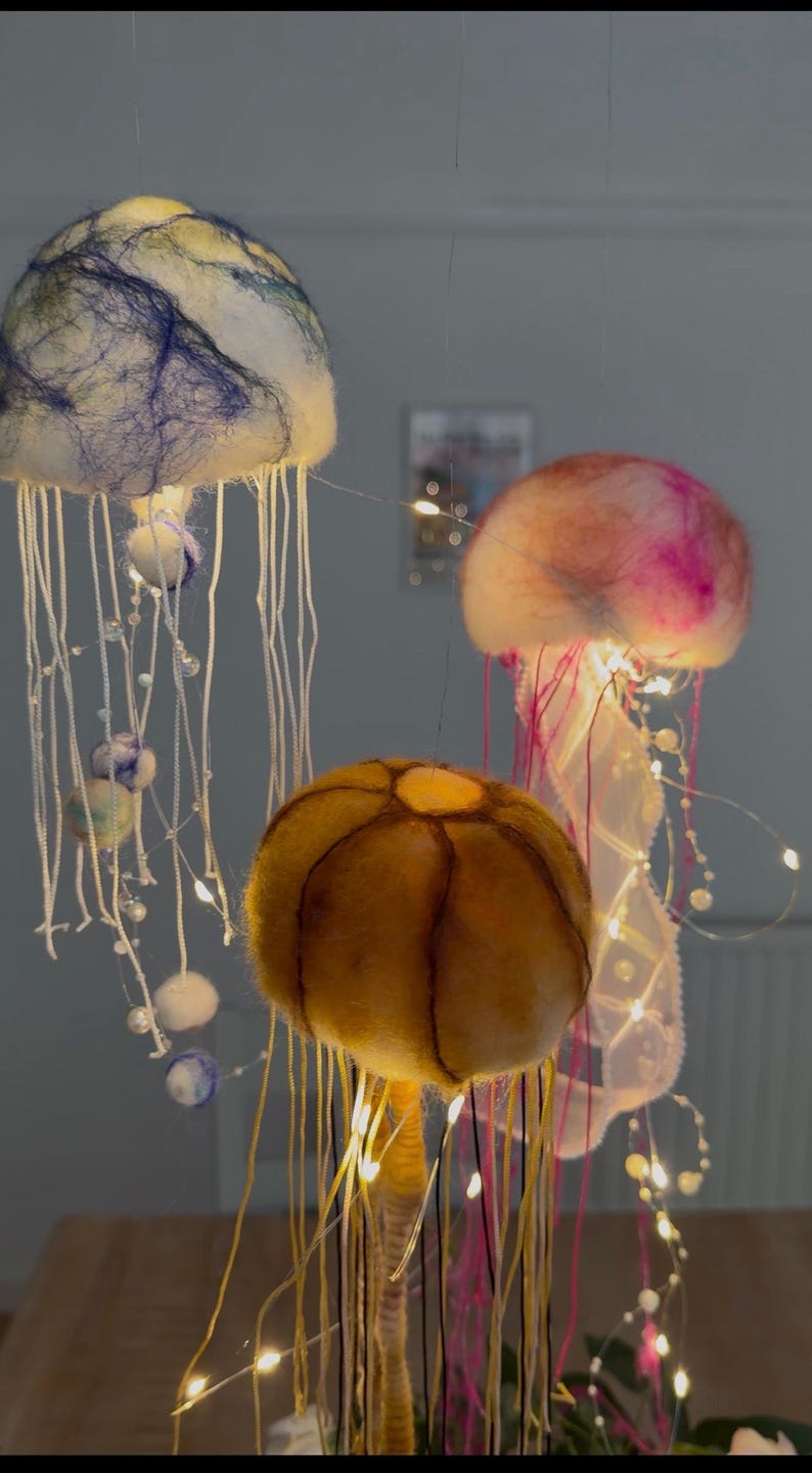 May include: Three hanging jellyfish decorations made of felted wool. One is white with blue fibers, one is pink and white, and one is golden yellow with dark brown segment lines. Each features long dangling white and colored threads and small LED lights.