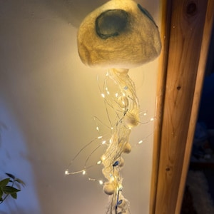 May include: A hanging jellyfish-shaped light fixture crafted from off-white felted wool. The dome features soft blue circular accents, while long, trailing tentacles made of white yarn and string lights emit a warm, glowing illumination.