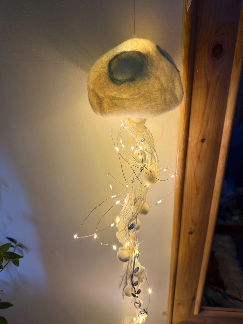 May include: A hanging jellyfish-shaped light fixture crafted from off-white felted wool. The dome features soft blue circular accents, while long, trailing tentacles made of white yarn and string lights emit a warm, glowing illumination.