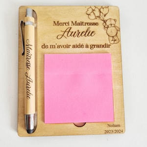May include: Wooden desk organiser with a pen holder, a sticky note pad, and a personalised message that reads "Merci Maitresse Aurelie de m'avoir aide a grandir". The message is engraved on the wood and includes a small cartoon bear holding balloons. The bottom of the organiser has the name "Noham" and the year "2023/2024" engraved on it.