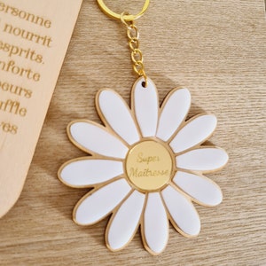 Op de afbeelding: Een gouden sleutelhanger met een witte madeliefje-bedel. De madeliefje heeft een gouden hart met de tekst "Super Maitresse" in goud.