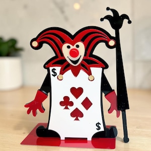 May include: A decorative card holder in the shape of a jester. The jester has a red and black hat, a white face with a red nose, and red gloves. The card displays card suit symbols and dollar signs.