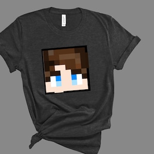 Puede incluir: Camiseta gris oscuro con un rostro pixelado con cabello castaño y ojos azules. El gráfico está enmarcado en un cuadrado negro. La camiseta es de material suave y tiene mangas enrolladas.