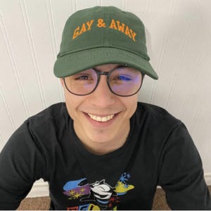 May include: A green baseball cap with the text "GAY & AWAY" in orange lettering. The person is wearing black-framed glasses and a black long-sleeve shirt with a graphic design.