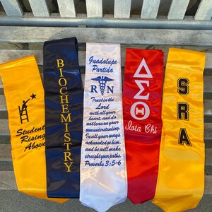 May include: Five graduation stoles in yellow, navy blue, white, and red. The stoles feature text and graphics, including "Student Rising Above," "BIOCHEMISTRY," "Guadalupe Partida," and Greek letters. The stoles are made of a shiny fabric.