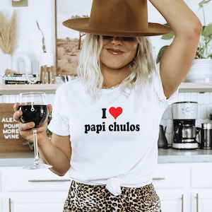 Puede incluir: Camiseta blanca con un corazón rojo y el texto "I love papi chulos" impreso en ella. La persona lleva un sombrero marrón y sostiene una copa de vino tinto.