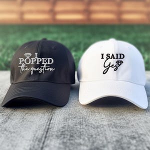 May include: Two baseball caps, one black and one white, with embroidered text. The black cap says "I popped the question" with a diamond symbol. The white cap says "I said Yes" with a diamond symbol.