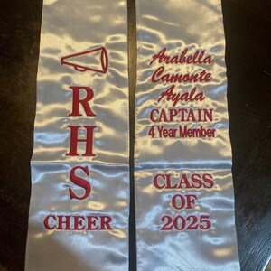 May include: Silver satin graduation sash with red embroidered text and a megaphone graphic. The sash reads "RHS CHEER" and "Arabella Camonte Ayala CAPTAIN 4 Year Member CLASS OF 2025".