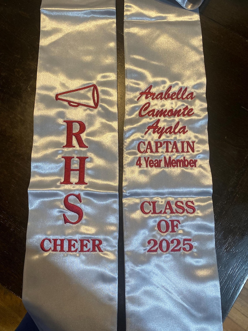 May include: Silver satin graduation sash with red embroidered text and a megaphone graphic. The sash reads "RHS CHEER" and "Arabella Camonte Ayala CAPTAIN 4 Year Member CLASS OF 2025".