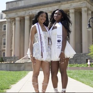 May include: Two women in white dresses and graduation stoles pose outside. One dress is floral, the other plain. The stoles read "PSYCHOLOGY" and "Sage A. Denise, Class of 2026". They smile in front of a large building with columns.