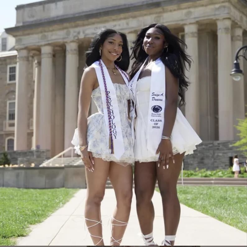 May include: Two women in white dresses and graduation stoles pose outside. One dress is floral, the other plain. The stoles read "PSYCHOLOGY" and "Sage A. Denise, Class of 2026". They smile in front of a large building with columns.