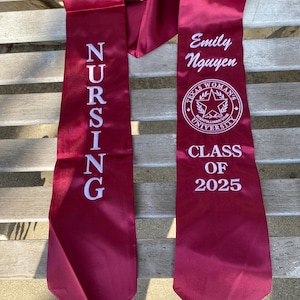 May include: Burgundy satin graduation sash with white embroidered text. One side reads "NURSING" vertically. The other side displays "Emily Nguyen", a university emblem, and "CLASS OF 2025".