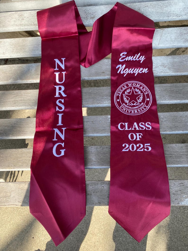 May include: Burgundy satin graduation sash with white embroidered text. One side reads "NURSING" vertically. The other side displays "Emily Nguyen", a university emblem, and "CLASS OF 2025".