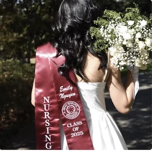 May include: A graduate wearing a claret nursing sash with the name "Emily Nguyen" and "Class of 2025" printed on it. The sash is over a white dress. The graduate is holding a bouquet of white flowers.