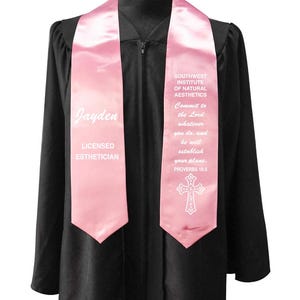 May include: A pink graduation stole with white text. The stole says "Jayden" and "LICENSED ESTHETICIAN" on one side. The other side says "SOUTHWEST INSTITUTE OF NATURAL AESTHETICS" and a bible verse. A white cross is at the bottom.