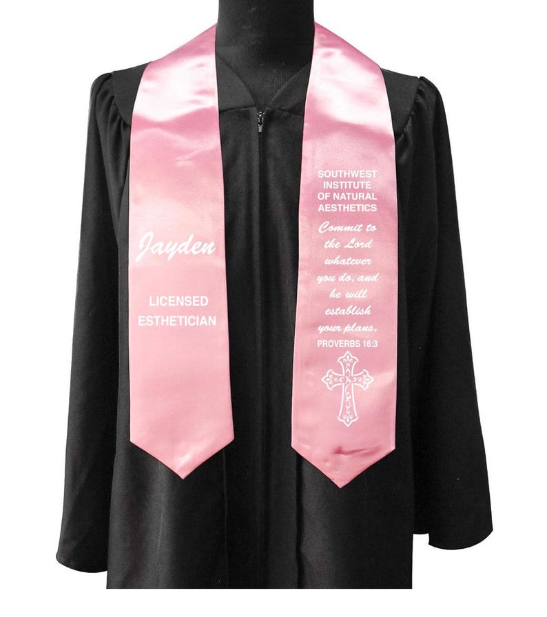 May include: A pink graduation stole with white text. The stole says "Jayden" and "LICENSED ESTHETICIAN" on one side. The other side says "SOUTHWEST INSTITUTE OF NATURAL AESTHETICS" and a bible verse. A white cross is at the bottom.