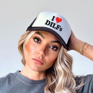 Puede incluir: Una gorra de camionero negra y blanca con un corazón rojo y el texto "I ♥ DILFS" en negro.