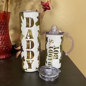 May include: Two white tumblers with lids. The larger tumbler has the word "DADDY" in a camouflage pattern. The smaller tumbler has the words "DADDY'S BOY" in a camouflage pattern.