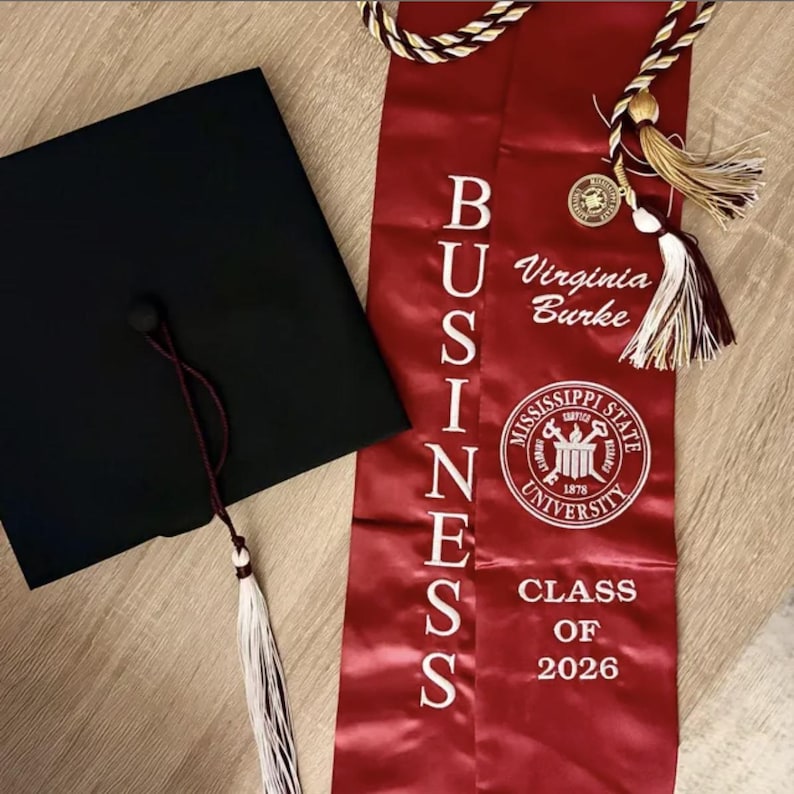 May include: A black graduation cap with a maroon tassel and a red satin sash. The sash reads "BUSINESS" vertically in white letters, with "Virginia Burke", the Mississippi State University seal, and "CLASS OF 2026".