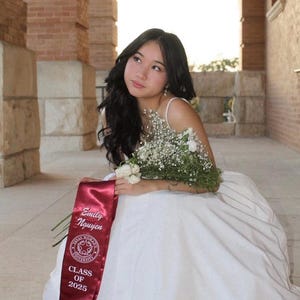 May include: A young woman in a white dress, holding a bouquet of white flowers and greenery, and wearing a maroon sash that reads "Emily Nguyen" and "Class of 2025". The background features brick columns and natural light.