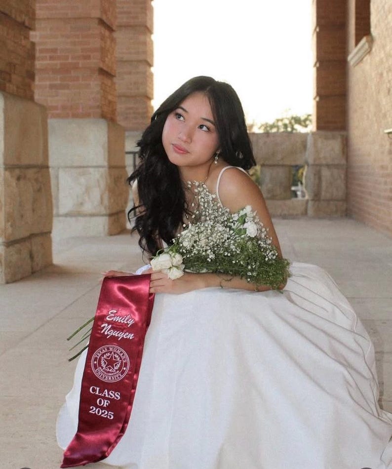 May include: A young woman in a white dress, holding a bouquet of white flowers and greenery, and wearing a maroon sash that reads "Emily Nguyen" and "Class of 2025". The background features brick columns and natural light.