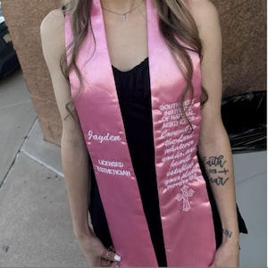 May include: A pink graduation sash with white embroidered text. The sash reads "Jayden" and "LICENSED ESTHETICIAN" on one side. The other side has text that reads "SOUTH EAST INSTITUTE OF NATURAL AESTHETICS". A cross is also embroidered on the sash.