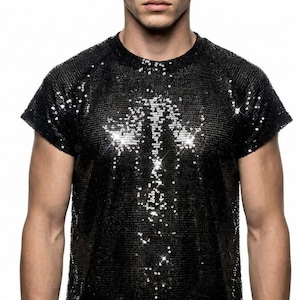 Op de afbeelding: Zwart pailletten T-shirt met korte mouwen. Het shirt is bedekt met kleine, glanzende pailletten die het licht reflecteren en een sprankelend effect creëren. Het T-shirt heeft een ronde hals en een getailleerd silhouet. Het model draagt het shirt tegen een witte achtergrond.