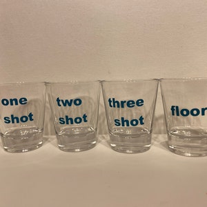 May include: A set of four clear glass shot glasses arranged in a row. Each glass features blue text printed on the front. The glasses are labelled one shot, two shot, three shot, and floor, respectively, in a simple, bold, sans-serif typeface.