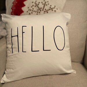 May include: A square white throw cushion featuring the word HELLO printed in large, thin, black, hand-drawn style capital letters centred on the front. The fabric appears smooth and matte, with the black text standing out against the white background.