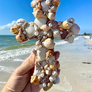 May include: A decorative cross crafted from various seashells and faux pearls. The cross features a mix of beige, brown, and white shells, with the pearls adding a touch of elegance. The cross is held against a backdrop of a beach and blue sky.