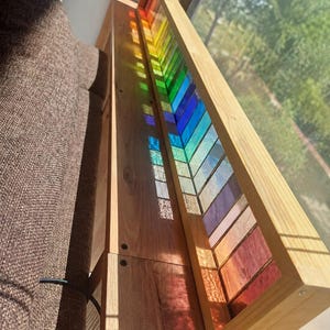 Peut inclure: Un long cadre rectangulaire en bois contenant une série de panneaux verticaux en verre teinté multicolore disposés en spectre. Les pièces de verre présentent des nuances de rouge, orange, jaune, vert, bleu et violet, projetant des reflets lumineux.