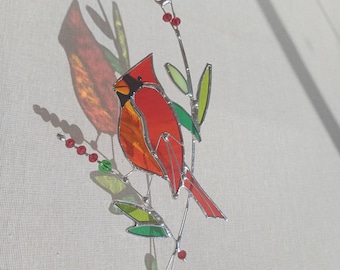 May include: A stained glass cardinal ornament with red, orange, and yellow glass, perched on a branch with green leaves and red berries. The ornament is hanging from a red ribbon.