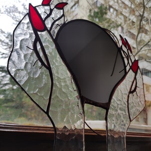 May include: Stained glass art piece featuring hands with red fingernails holding a dark gray oval. The glass has a textured, clear appearance with dark brown outlines. The artwork is displayed in front of a window.
