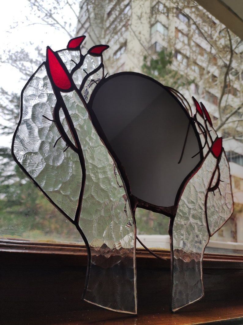 May include: Stained glass art piece featuring hands with red fingernails holding a dark gray oval. The glass has a textured, clear appearance with dark brown outlines. The artwork is displayed in front of a window.