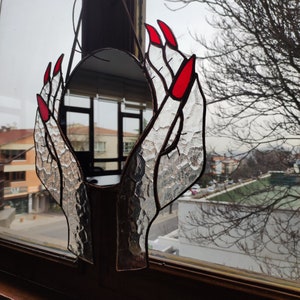 May include: Stained glass mirror in the shape of hands with red fingernails. The hands are holding a round mirror. The glass has a textured appearance. The mirror is hanging in front of a window.